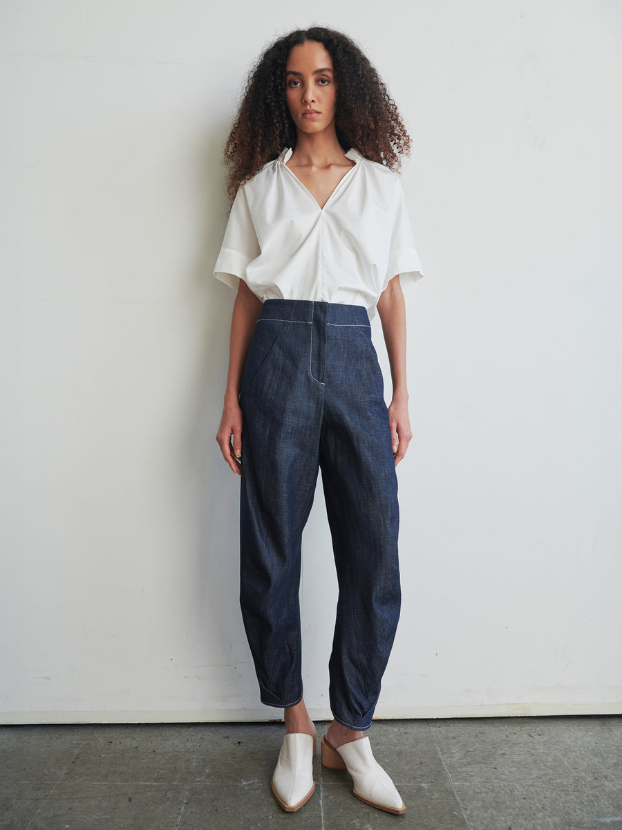 A person with long, curly hair stands against a plain white wall, wearing a loose white collared shirt and the high-waisted Takeo Pant from Zero + Maria Cornejo, made from 100% organic cotton. White pointed shoes complete their effortlessly chic ensemble.