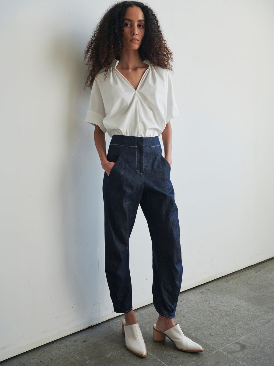 A person with long curly hair stands against a white wall, sporting a white short-sleeved shirt and the Takeo Pant, crafted by Zero + Maria Cornejo from 100% organic cotton. With hands in pockets and wearing white heeled shoes, they gaze confidently towards the camera.