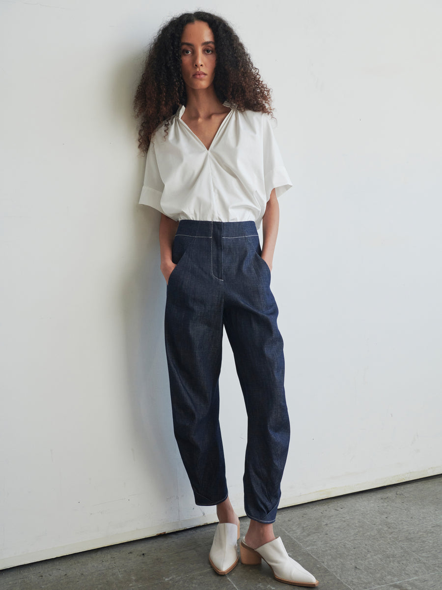 A person with curly hair stands against a white wall, wearing a white blouse and the high-waisted Takeo Pant by Zero + Maria Cornejo, crafted from 100% organic cotton. Their look is completed with white shoes, and they maintain a neutral expression with their hands resting in their pockets.