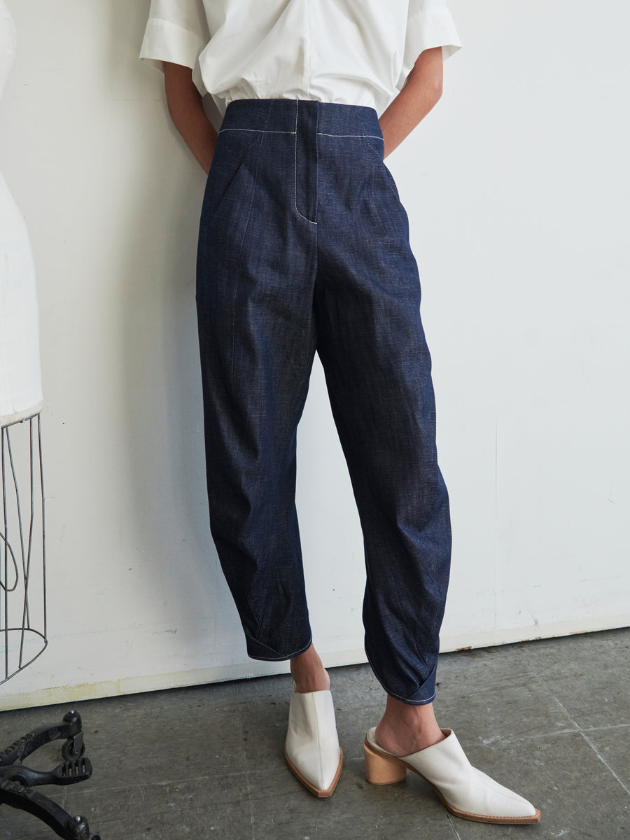 A person stands against a white wall wearing a white shirt and the Takeo Pant, crafted from dark blue organic denim by Zero + Maria Cornejo. They have one hand in their pocket and are wearing medium-heeled, white slip-on shoes on a concrete floor. A mannequin is partially visible on the side.