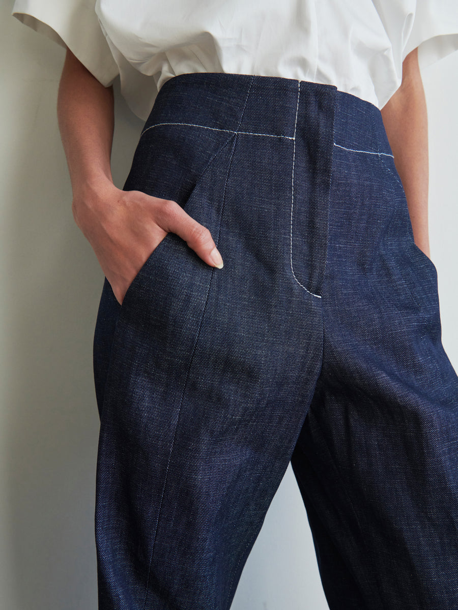 A person wearing a white shirt and the Takeo Pant in dark blue, by Zero + Maria Cornejo, featuring high-waisted organic denim with hands in pockets. The fabric of the pants highlights visible stitching and a subtle texture, crafted from GOTS-certified cotton for an environmentally friendly choice.