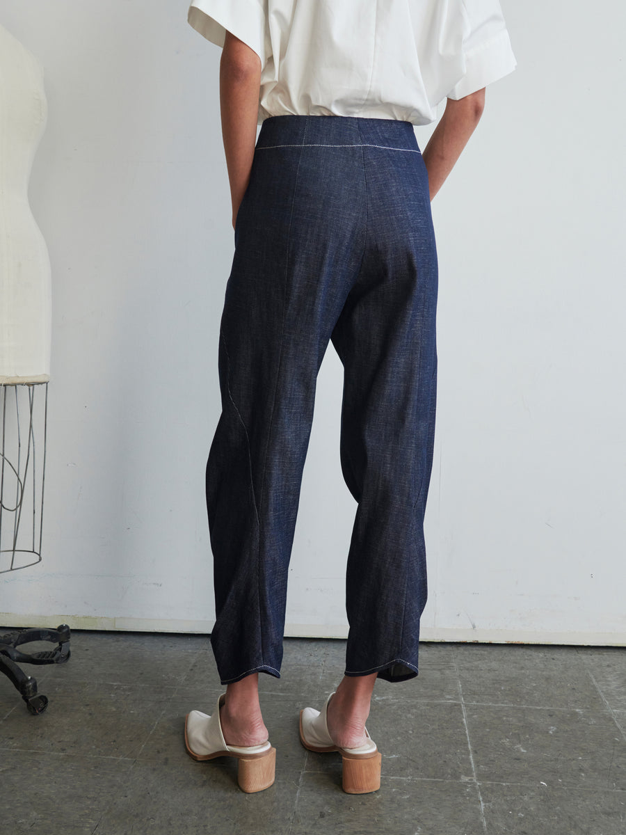 A person in Takeo Pants by Zero + Maria Cornejo and white shoes stands facing away. They are wearing a white top made from GOTS-certified cotton, and the room has a minimalist style with a mannequin visible on the left.