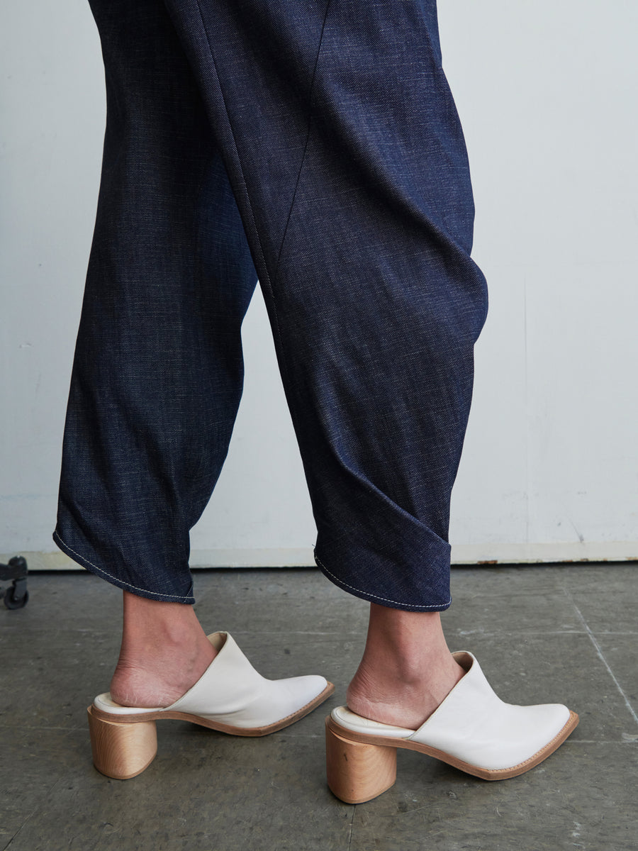 A person wearing the Takeo Pant by Zero + Maria Cornejo in organic navy blue denim and white mules with wooden block heels stands on a concrete floor against a plain light-colored wall. The focus is on their legs and shoes.