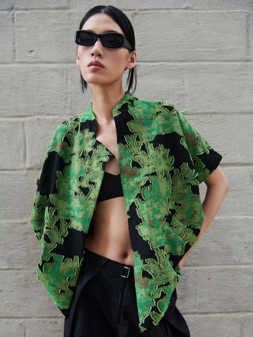 A person wearing black rectangular sunglasses, a green and black floral patterned open Ari Shrug by Zero + Maria Cornejo, and black high-waisted pants stands in front of a light gray brick wall. The shrug, crafted from organic cotton, is slightly open, revealing a black top underneath.
