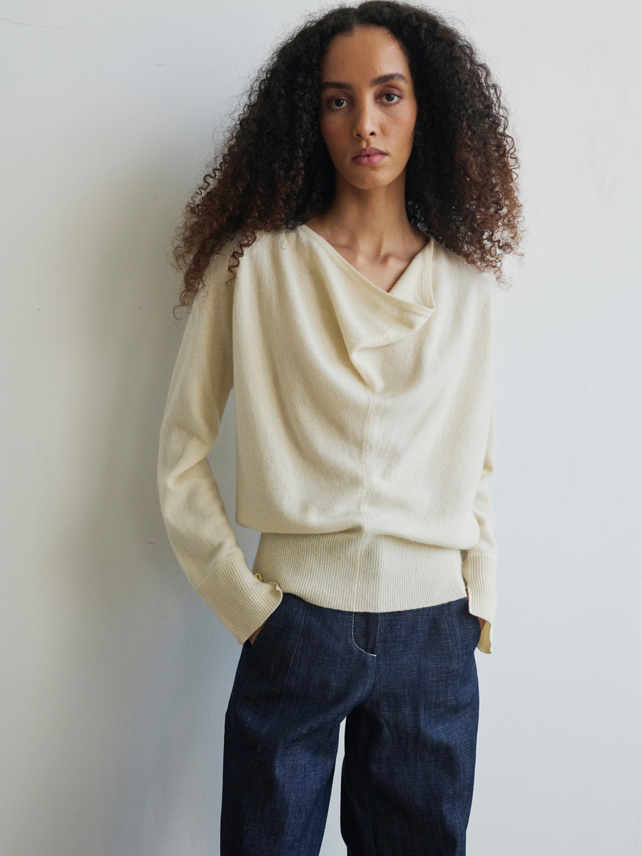 A person with long curly hair stands against a plain wall, wearing the Knit Evi Top by Zero + Maria Cornejo, an eco-friendly sweater made from recycled cashmere, paired with dark pants. They have a calm expression and hands in their pockets.