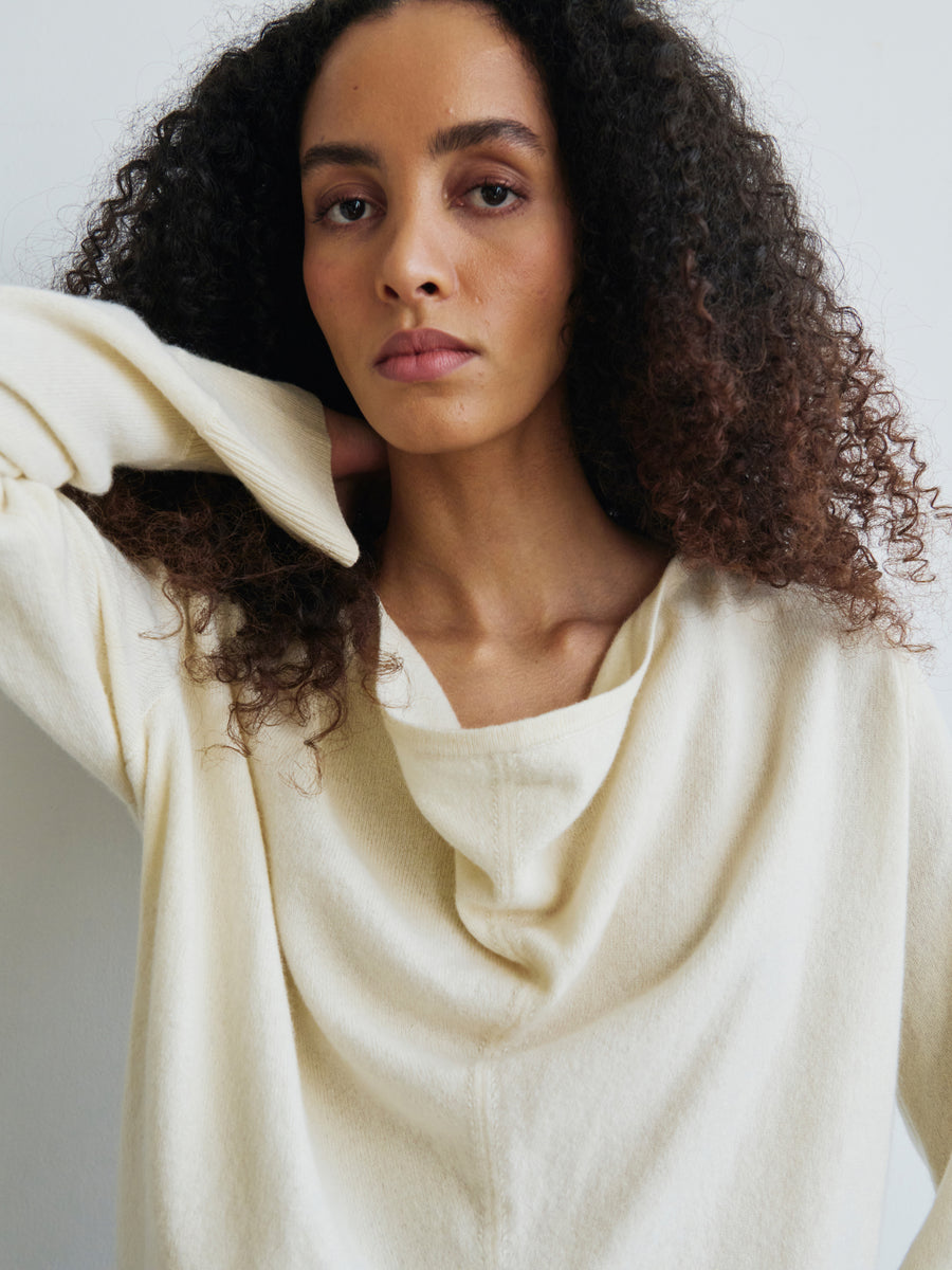 A person with curly hair poses against a light background, wearing the cream-colored Knit Evi Top, a long-sleeve sweater by Zero + Maria Cornejo. Crafted from recycled cashmere with a subtle cowl neck, their expression is calm and contemplative as one hand rests near their neck.