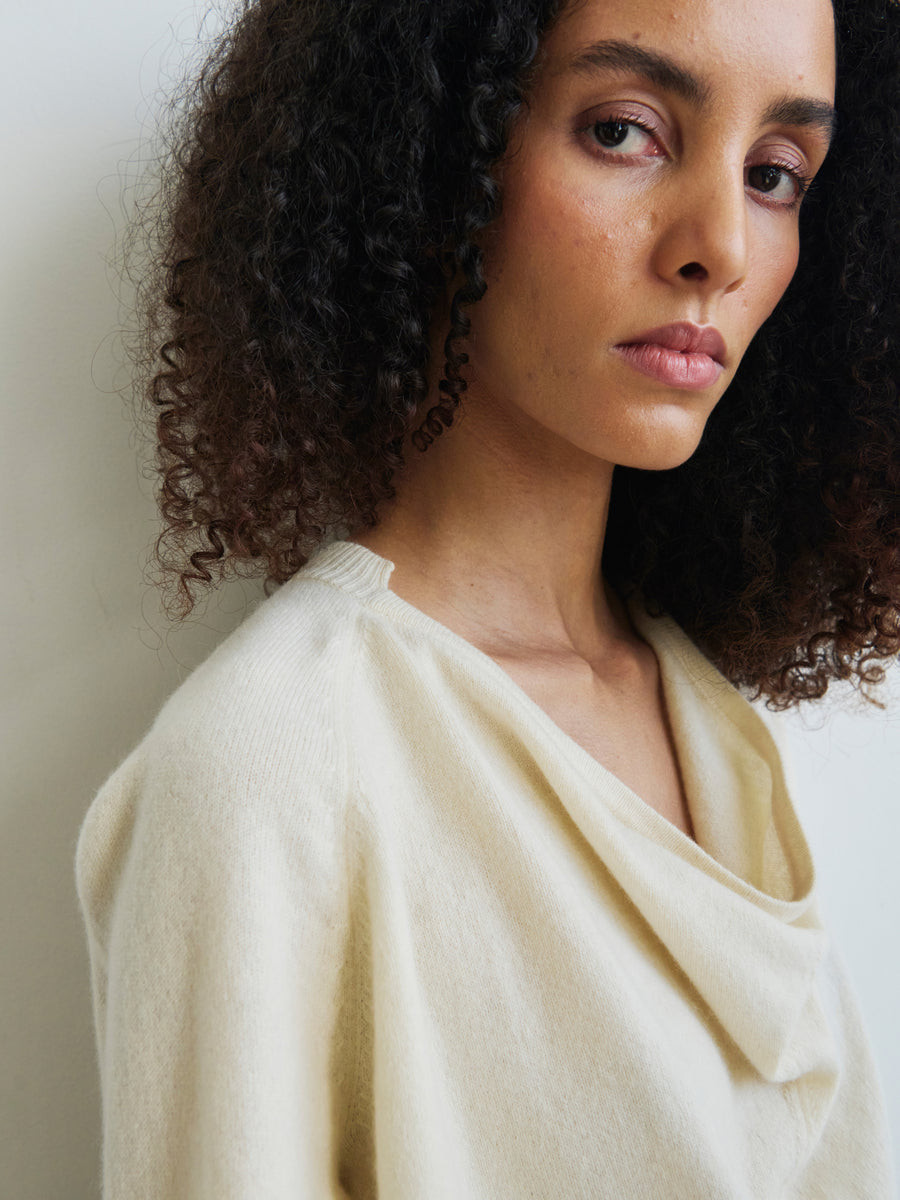 A person with curly hair is wearing the Knit Evi Top by Zero + Maria Cornejo, crafted from sustainably-sourced cashmere in a cream hue. They are positioned against a light background with a calm expression, gazing slightly off-camera, exuding a serene and contemplative vibe.