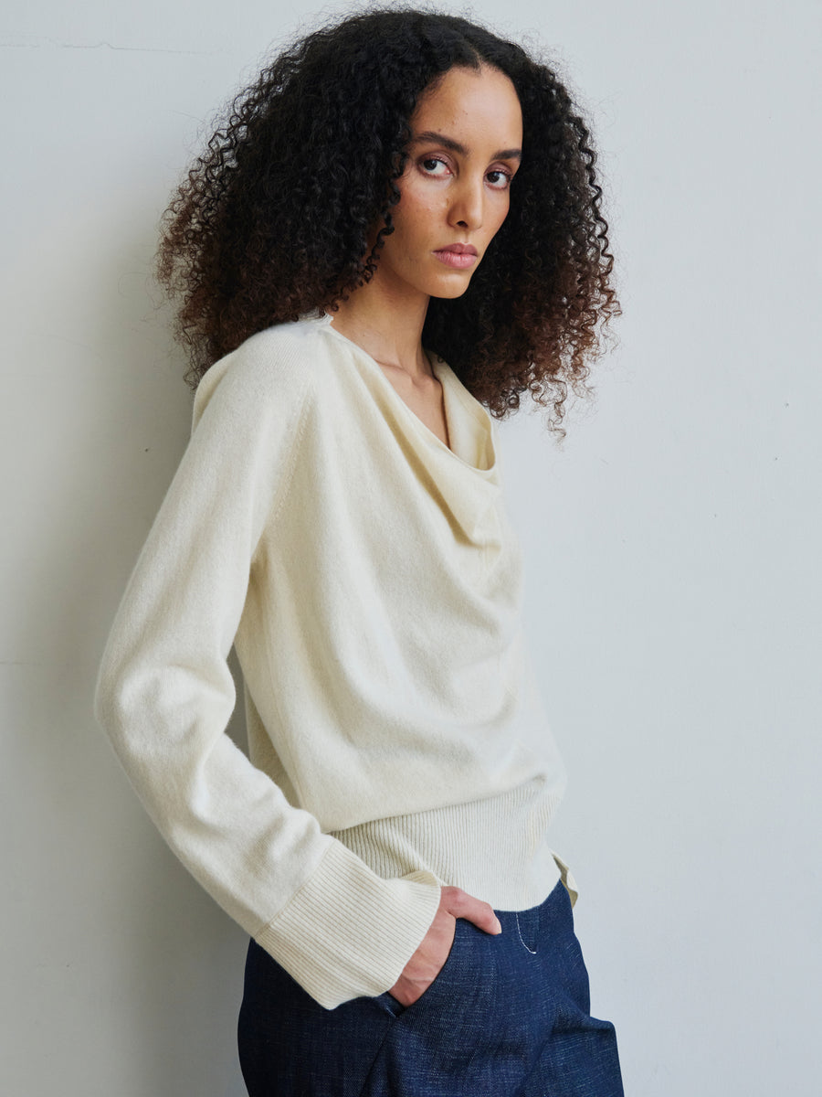 A person with curly hair, dressed in the Knit Evi Top by Zero + Maria Cornejo, paired with dark pants, stands against a plain light-colored wall. They gaze directly at the camera with a neutral expression.