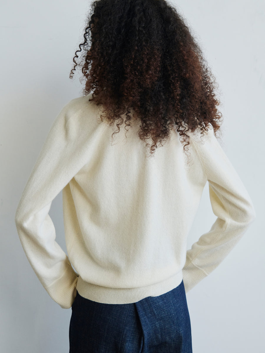 Wearing the Knit Evi Top by Zero + Maria Cornejo, a person with curly hair faces away, wrapped in an eco-friendly cream-colored sweater and dark pants, set against a light background.