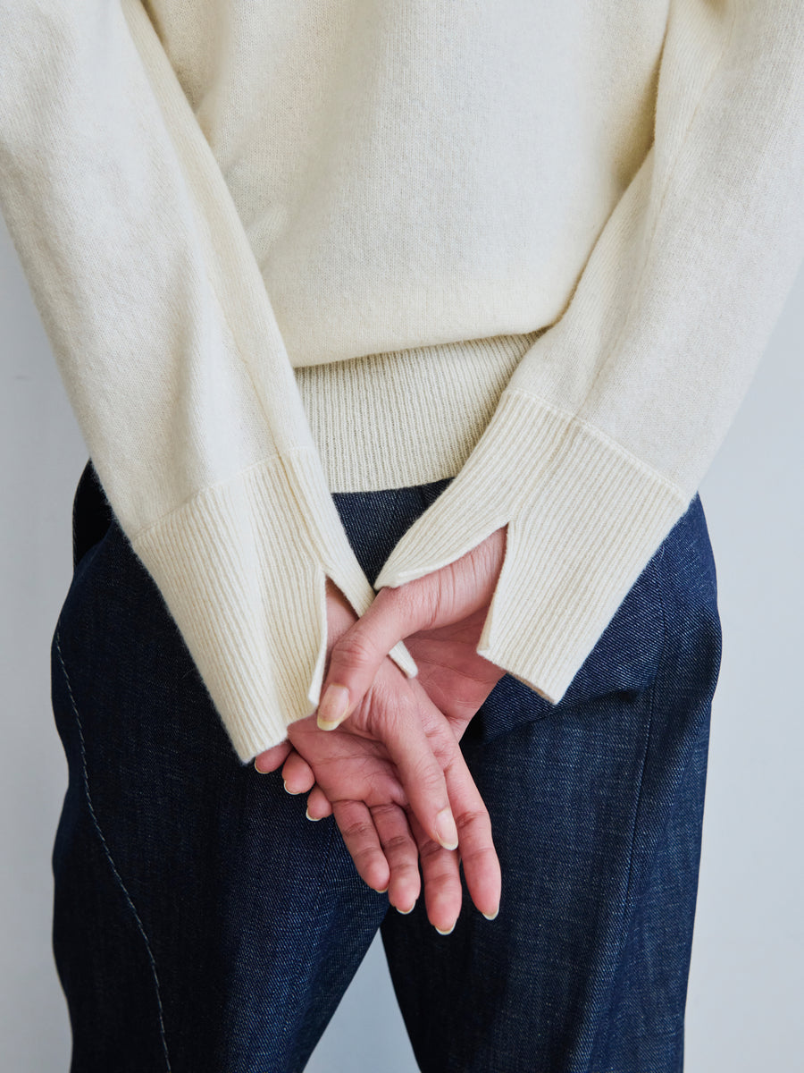 A person wearing the Knit Evi Top from Zero + Maria Cornejo stands with their hands clasped behind their back, showcasing a cream sweater made of recycled cashmere. The dark jeans complement the long sleeves with thumbholes, adding a touch of detail, while the sustainably-sourced cashmere emphasizes an eco-friendly option in a neutral setting.