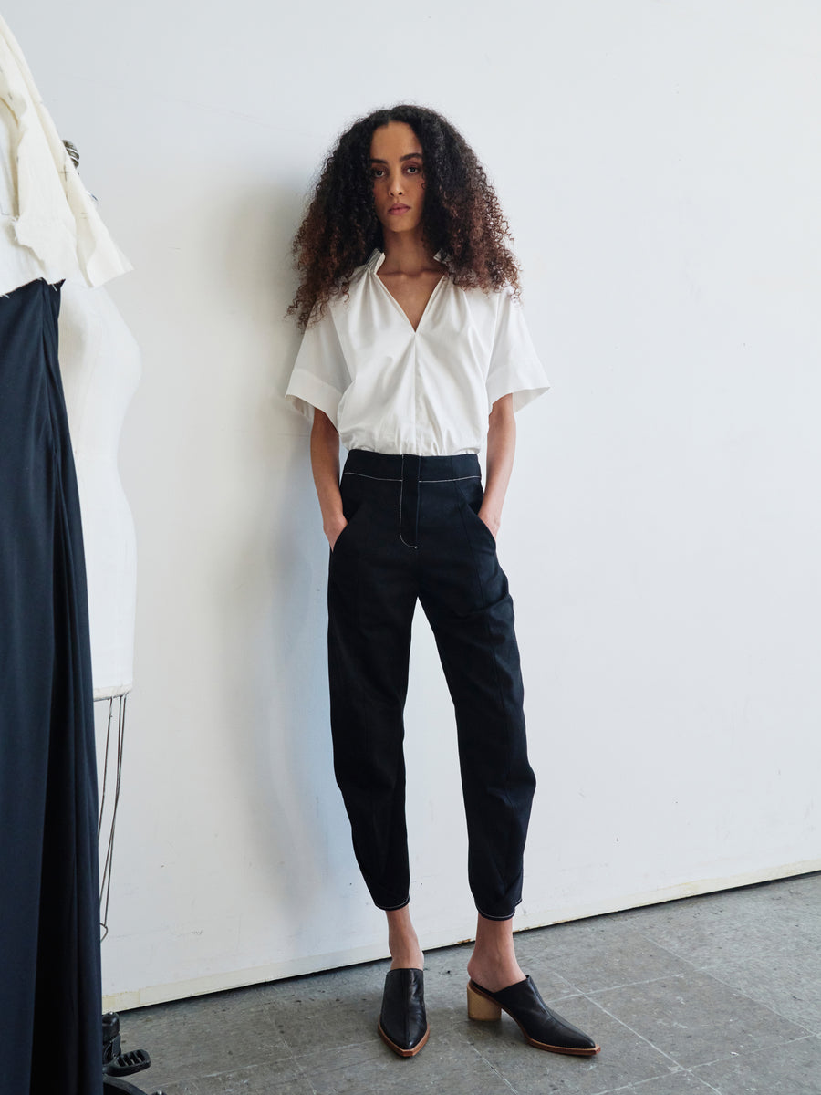 A person with curly hair stands confidently against a white wall, wearing a white blouse made from GOTS-certified cotton and the black organic denim Takeo Pant by Zero + Maria Cornejo. With their hands in their pockets, they exude a relaxed and stylish demeanor, completed with black slip-on shoes.