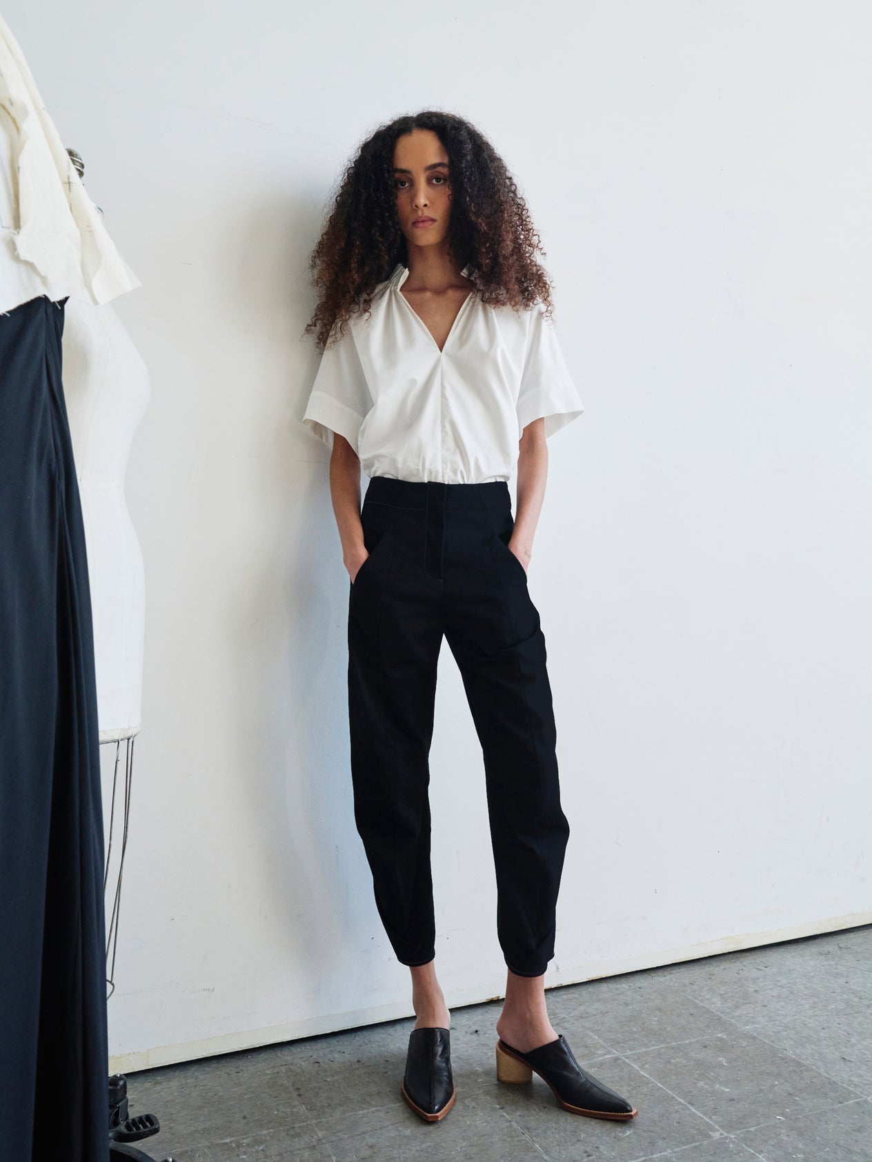 A person with curly hair stands against a white wall, wearing a white blouse and the high-waisted Takeo Pant by Zero + Maria Cornejo, crafted from 100% organic cotton. Their look is completed with white shoes, and they maintain a neutral expression with their hands resting in their pockets.