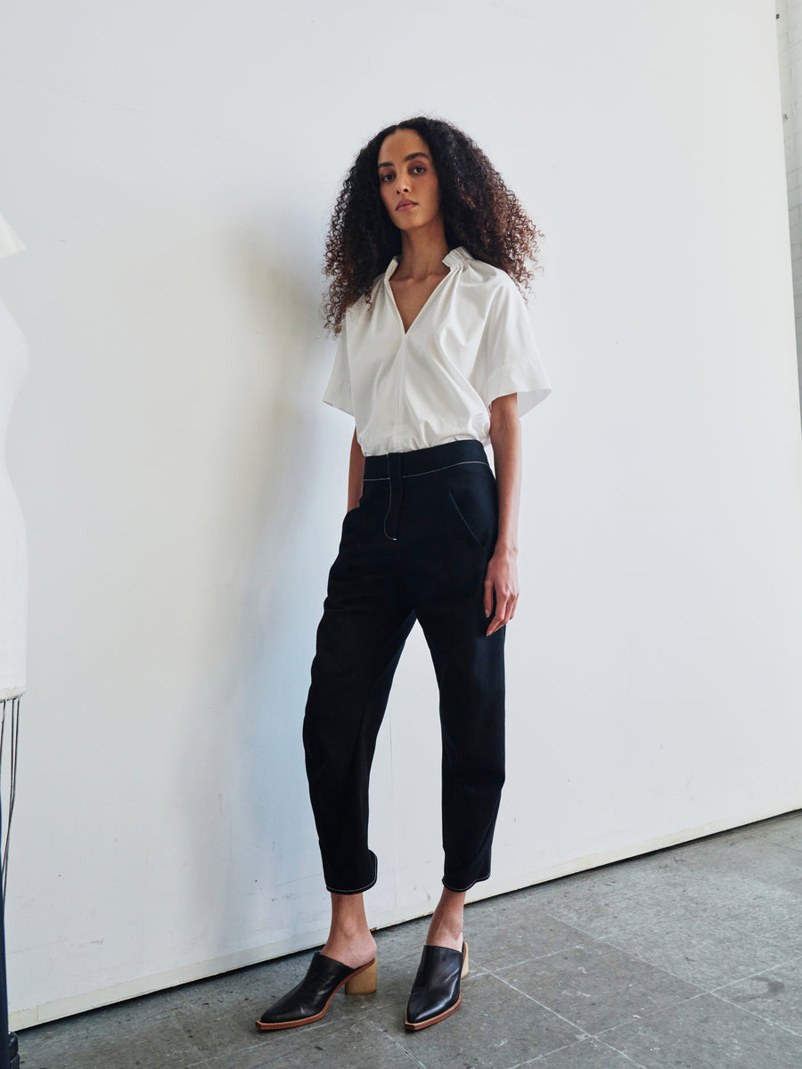 A person with curly hair stands against a plain white wall, dressed in a short-sleeved white shirt crafted from GOTS-certified cotton and high-waisted Takeo Pants from Zero + Maria Cornejo, made of organic black denim. Black slip-on shoes complete the look. The concrete floor adds an industrial touch, and a mannequin can be seen on the left side of the image.