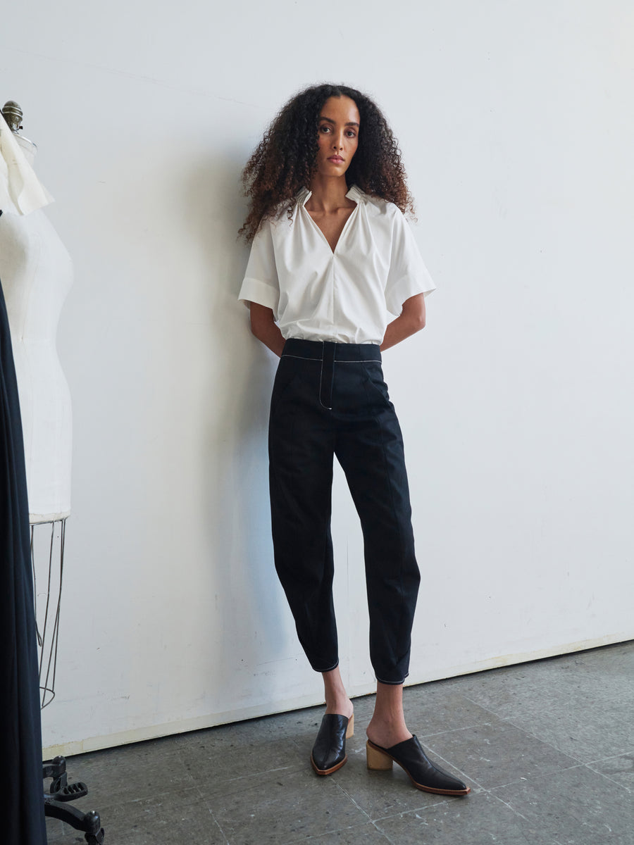 A person with curly hair stands against a white wall in a well-lit room. They are wearing a white short-sleeved shirt and the GOTS-certified Takeo Pant by Zero + Maria Cornejo, paired with black shoes. A dress form is partially visible on the left.