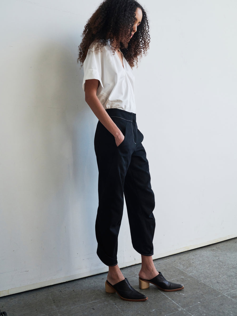 A person stands in a relaxed pose with hands in pockets, wearing a white blouse, the Takeo Pant by Zero + Maria Cornejo, and black heeled mules. The backdrop is a plain, light-colored wall.