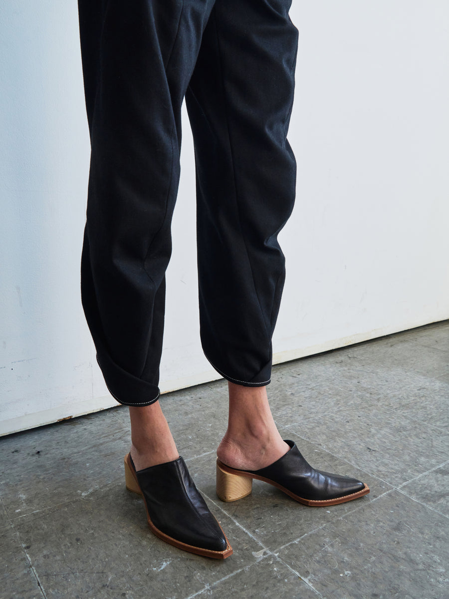 A person dressed in sleek Takeo Pants by Zero + Maria Cornejo and black heeled, open-back leather shoes stands on a gray tiled floor against a white wall.