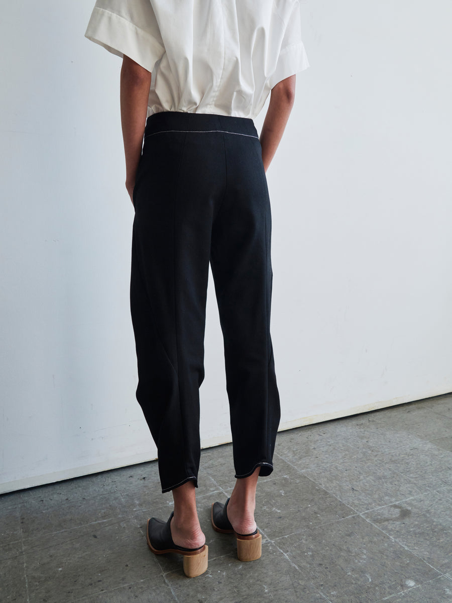 A person wearing a white short-sleeve shirt, Zero + Maria Cornejo's Takeo Pant in organic denim, and black clogs with wooden heels stands facing a wall in a minimalist room with a concrete floor. The image captures the back view from the shoulders down.