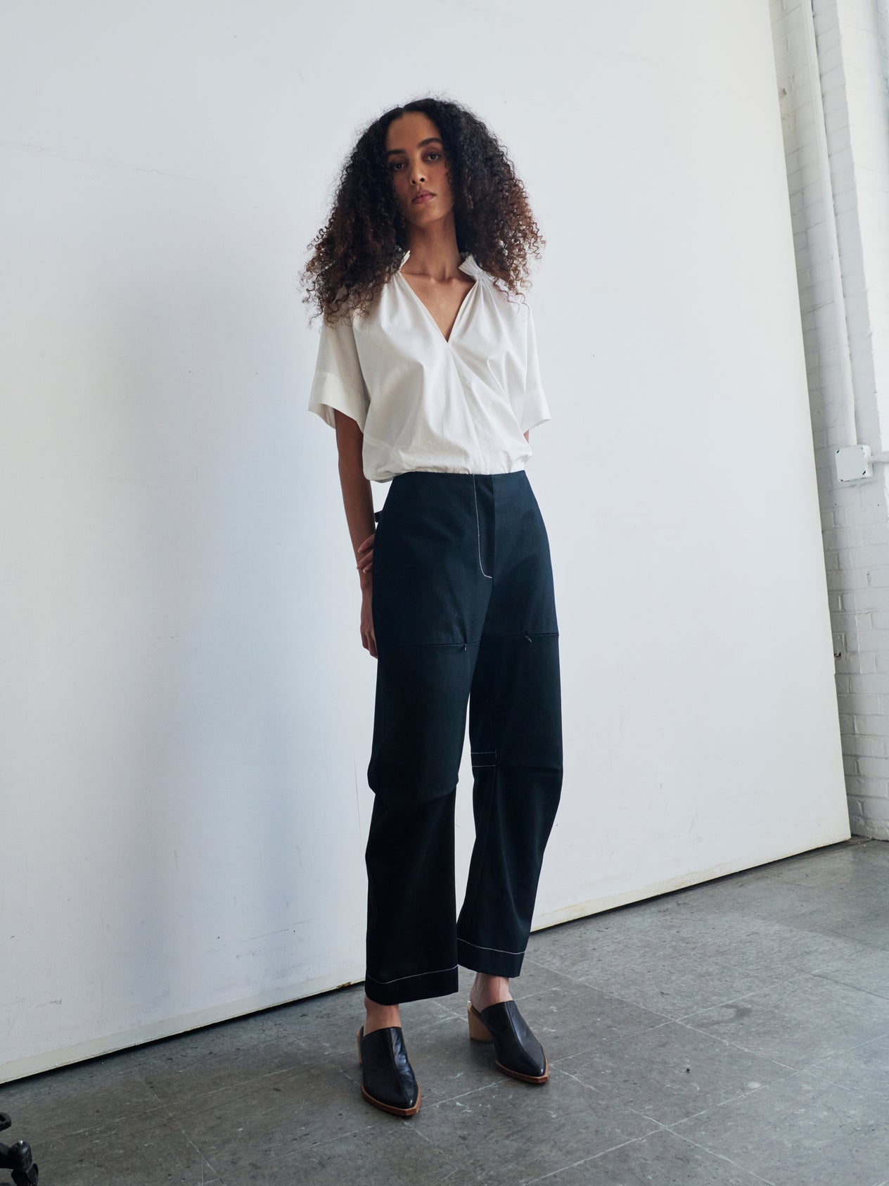 A confident person with curly hair poses against a white wall, wearing Zero + Maria Cornejo's Denim Biker Pant and white ankle boots. The cropped pant accentuates their stance as they rest one hand charmingly on their hair.