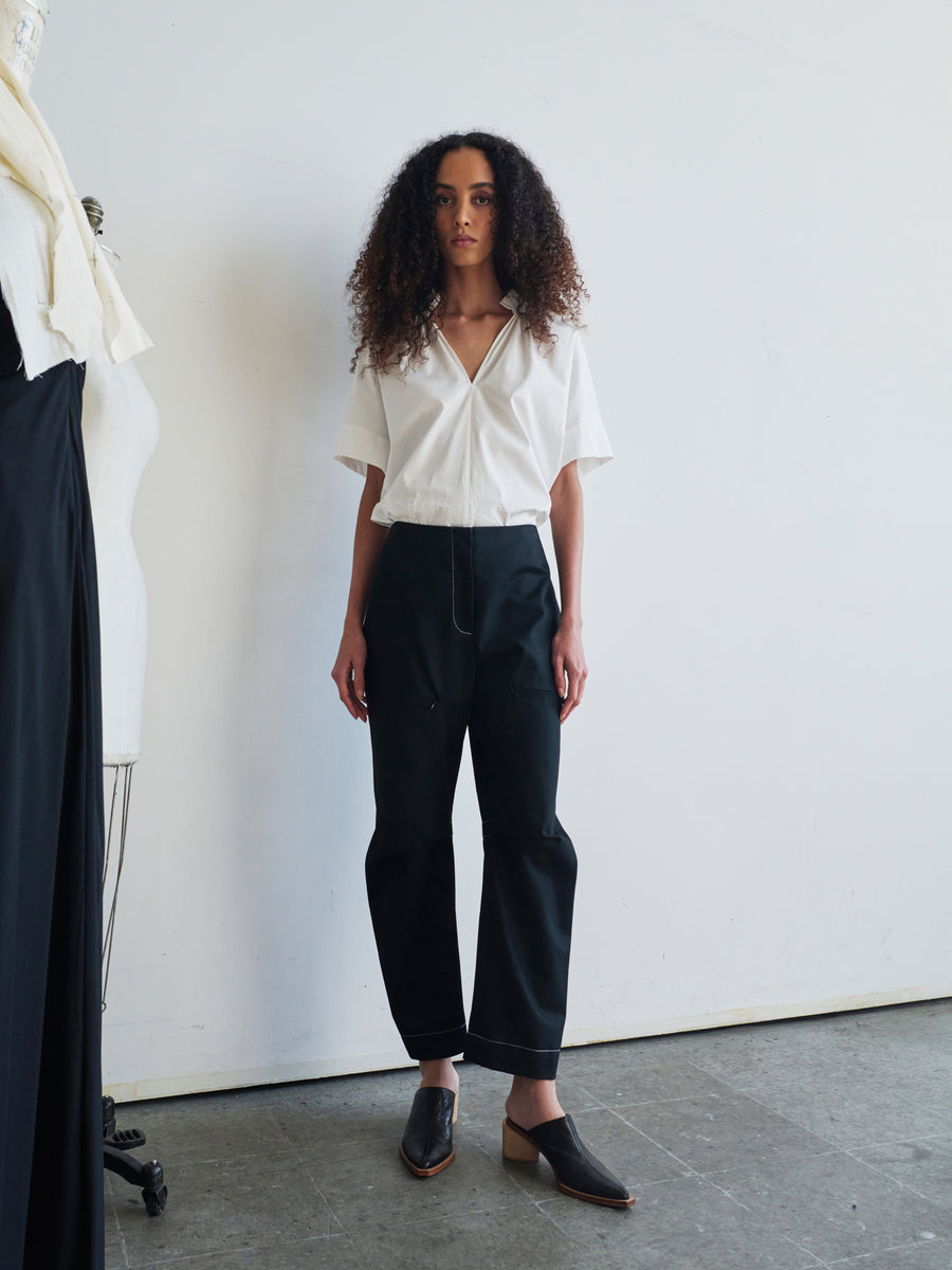 A person with curly hair stands against a white wall wearing a white shirt, the Denim Biker Pant by Zero + Maria Cornejo, and black slip-on shoes. On the left, there is a mannequin and fabric visible.