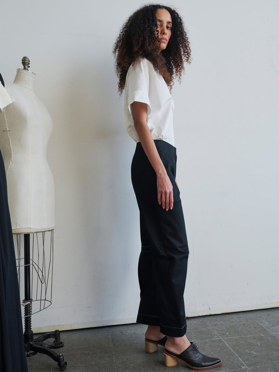 A curly-haired person stands in a minimalist room wearing a white shirt, Denim Biker Pants by Zero + Maria Cornejo, and brown heeled shoes. A tailor's mannequin is visible against the white wall in the background.