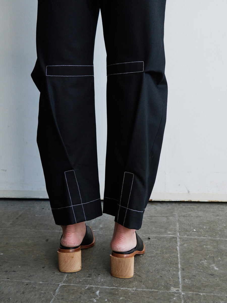 A person in Zero + Maria Cornejo's Denim Biker Pant, featuring cropped black design with white stitching and crafted from GOTS-certified cotton, pairs them with wooden heeled sandals on a tiled floor against a white wall background.