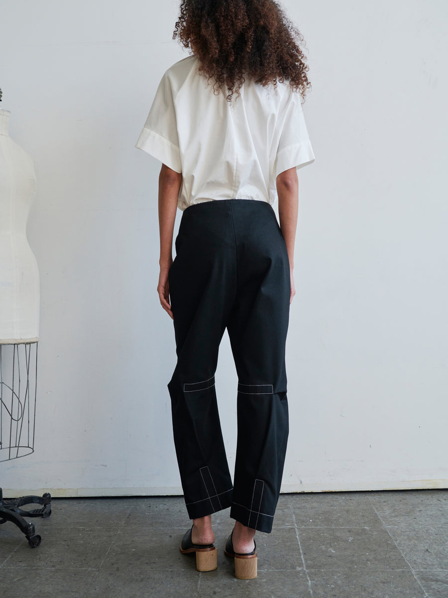 A person with curly hair stands against a white wall, wearing a white short-sleeve shirt and black Denim Biker Pant by Zero + Maria Cornejo, featuring stitching details. They sport open-toed sandals, with a part of a mannequin base visible on the left.