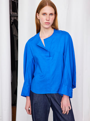 A woman with straight, light brown hair wears the Zero + Maria Cornejo Long-Sleeved Akeo Shirt—a bright blue, round-neck blouse—paired with dark pants, standing calmly before white curtains and looking at the camera with a neutral expression.