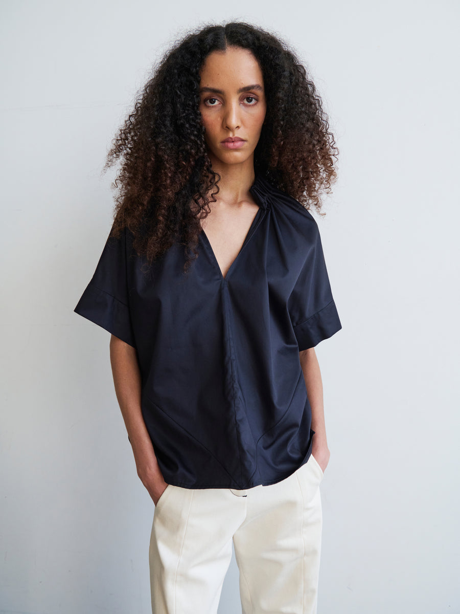 A person with long, curly hair wearing an oversized-fit Malia shirt from Zero + Maria Cornejo and light-colored pants stands against a plain white background with hands in pockets.