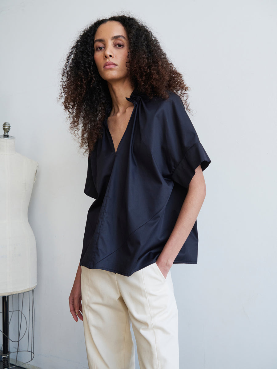 A person with curly hair wearing the loose black cotton Malia Shirt by Zero + Maria Cornejo and white pants stands next to a dress form against a plain background.