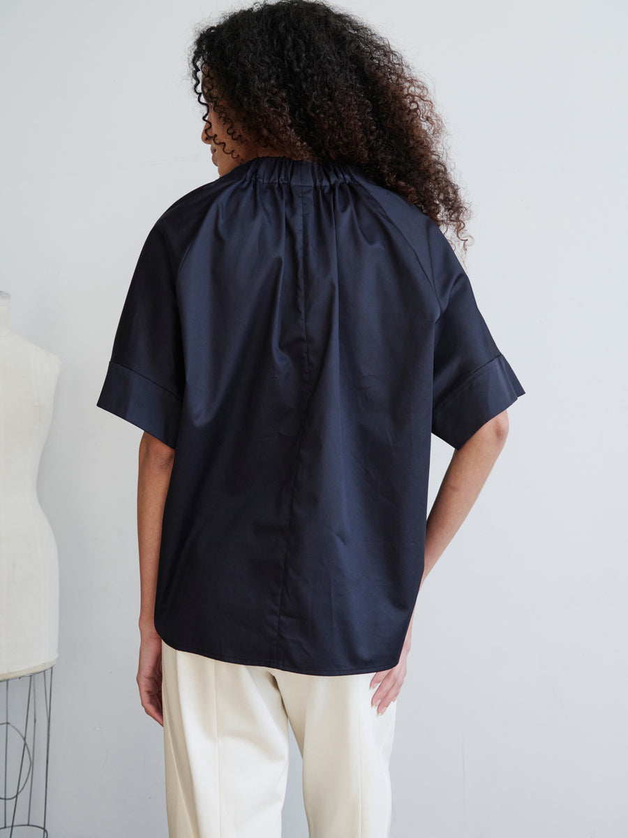 A person with curly hair stands with their back to the camera, dressed in an oversized fit Malia Shirt from Zero + Maria Cornejo and white pants. They are positioned in front of a light-colored wall, where part of a dress form is visible on the left.