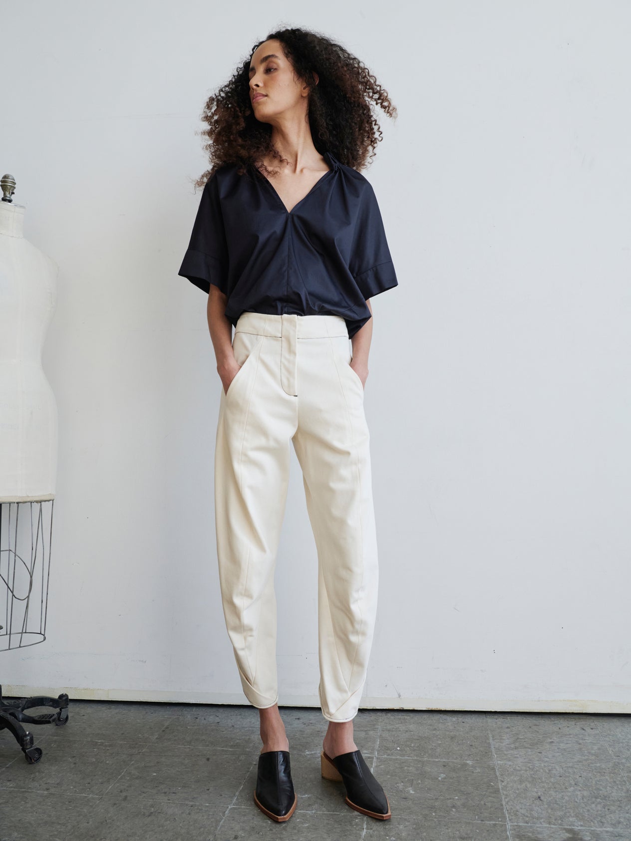 A person with curly hair stands against a white wall, wearing a white blouse and the high-waisted Takeo Pant by Zero + Maria Cornejo, crafted from 100% organic cotton. Their look is completed with white shoes, and they maintain a neutral expression with their hands resting in their pockets.
