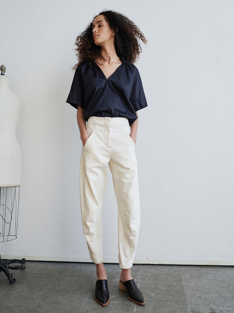 A person with curly hair stands confidently, hands in pockets, wearing a dark V-neck top and light-colored, high-waisted Takeo Pant by Zero + Maria Cornejo. A dress form is partially visible beside them against a plain wall.