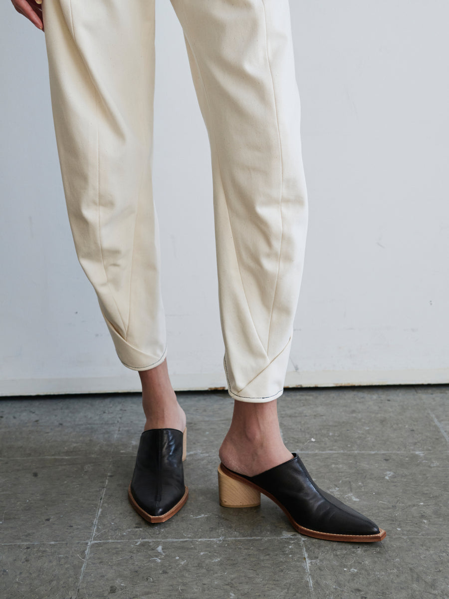 A person wearing Zero + Maria Cornejo's Takeo Pant made from GOTS-certified cotton and black heeled mules is standing on a gray floor. The mules feature pointed toes and wooden block heels, set against a plain, light-colored wall.