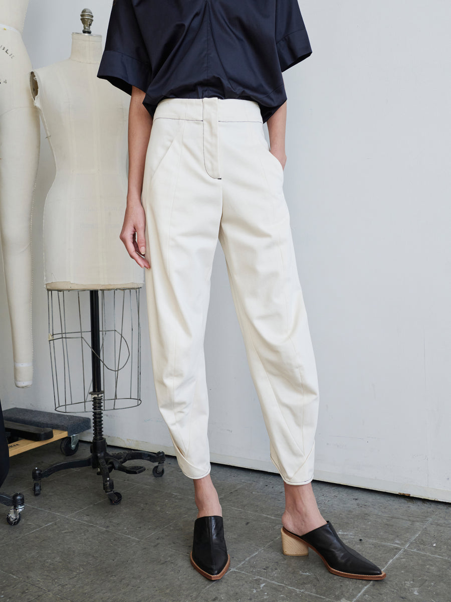 A person wearing the Takeo Pant in white by Zero + Maria Cornejo, which may be made from GOTS-certified cotton, pairs it with a dark shirt while standing beside dress forms. They complete their outfit with black slip-on shoes featuring a wooden heel, set against the backdrop of a fashion studio with a concrete floor.