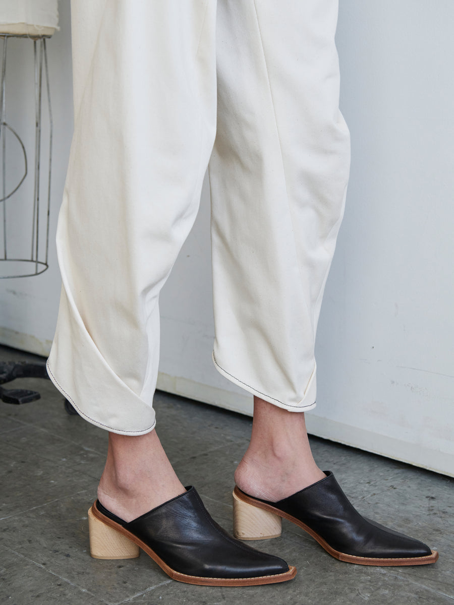 A person dressed in the Takeo Pant from Zero + Maria Cornejo, possibly crafted from GOTS-certified cotton, is positioned against a gray wall. They are wearing black leather mules featuring wooden block heels, and the concrete floor highlights their style. The base of a stool can be seen in the background.