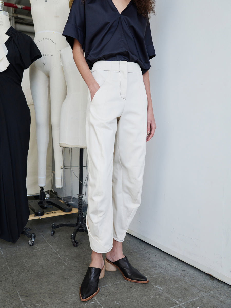 A person wearing a black blouse and the high-waisted, GOTS-certified Takeo Pant by Zero + Maria Cornejo stands with hands in pockets. They are wearing black slip-on shoes, and two dress forms are visible in the background on a concrete floor.