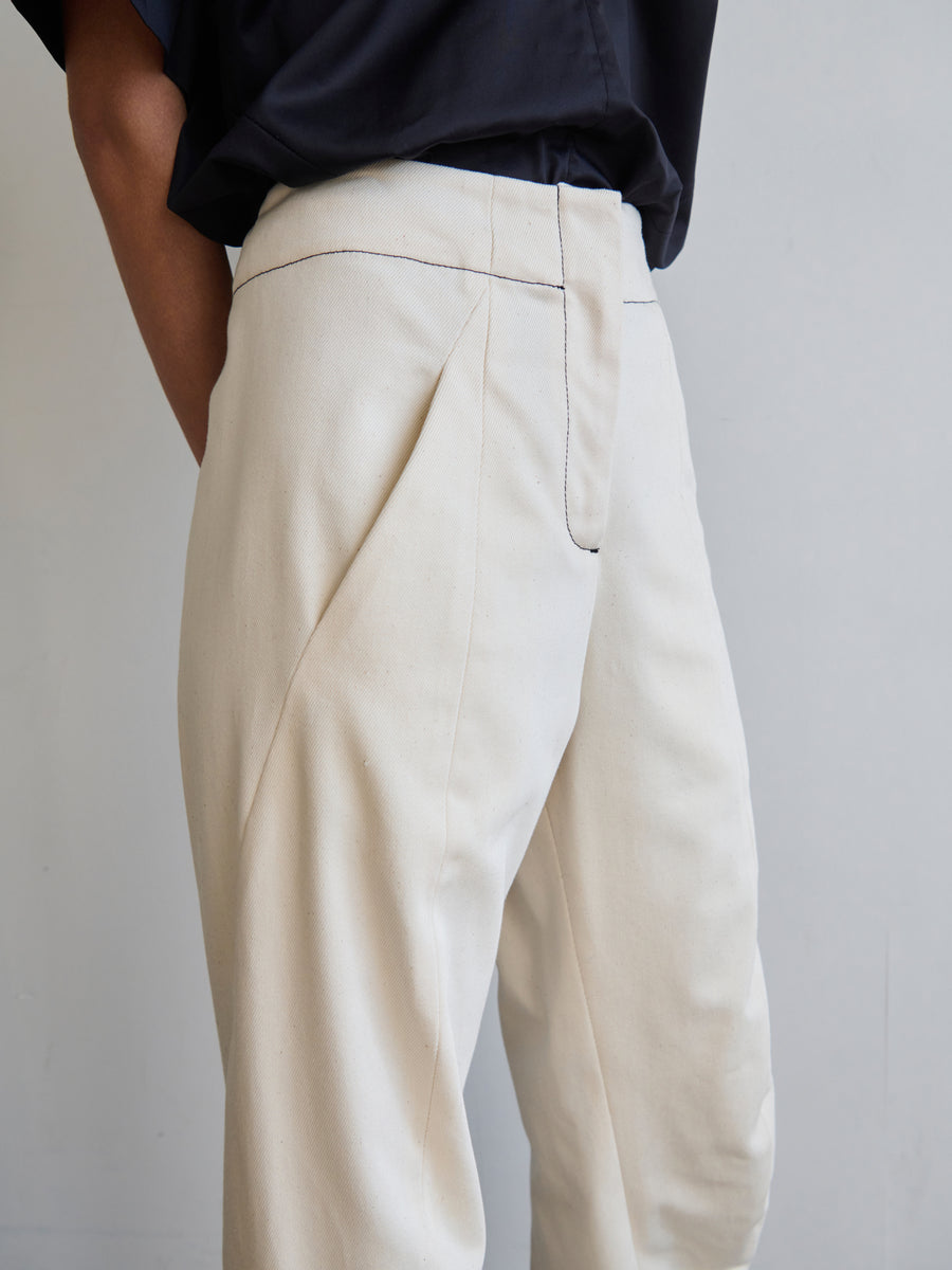 A person dressed in Zero + Maria Cornejo's Takeo Pant, which features a light beige color, high waist, and pleats crafted from GOTS-certified cotton, stands against a plain background. The upper portion of their dark shirt is partially visible as they relax with their hands tucked into the pant pockets.