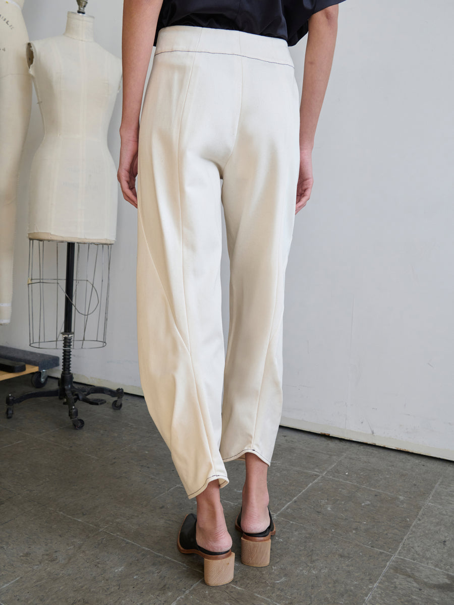 A person wearing Zero + Maria Cornejo's Takeo Pant in a loose-fitting cream style, crafted from Cradle 2 Cradle Certified™ Gold fabric, and paired with black sandals stands with their back to the camera. Mannequins are visible to the side in a workshop setting.