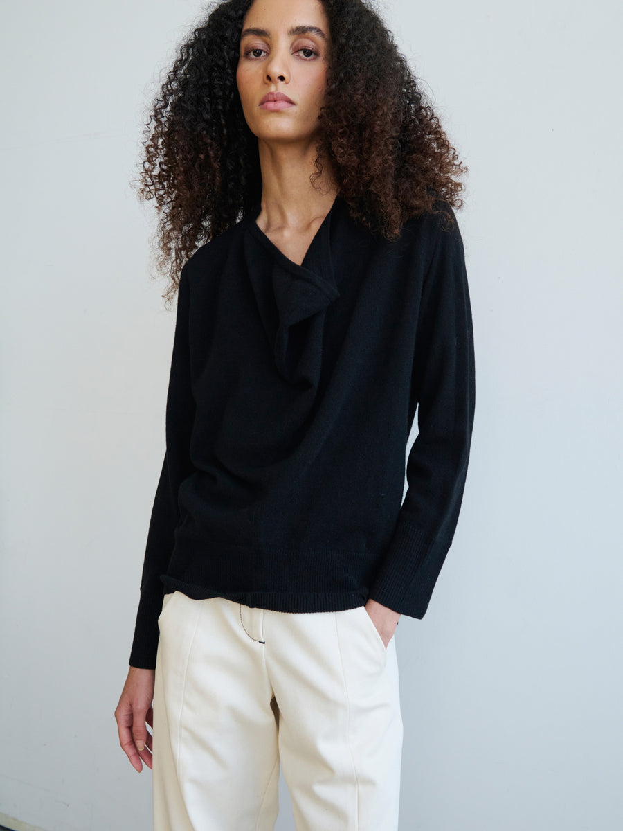 A person with curly hair is wearing the Knit Evi Top by Zero + Maria Cornejo, a black long-sleeved top crafted from eco-friendly knitwear, paired with light-colored pants, standing against a plain background.