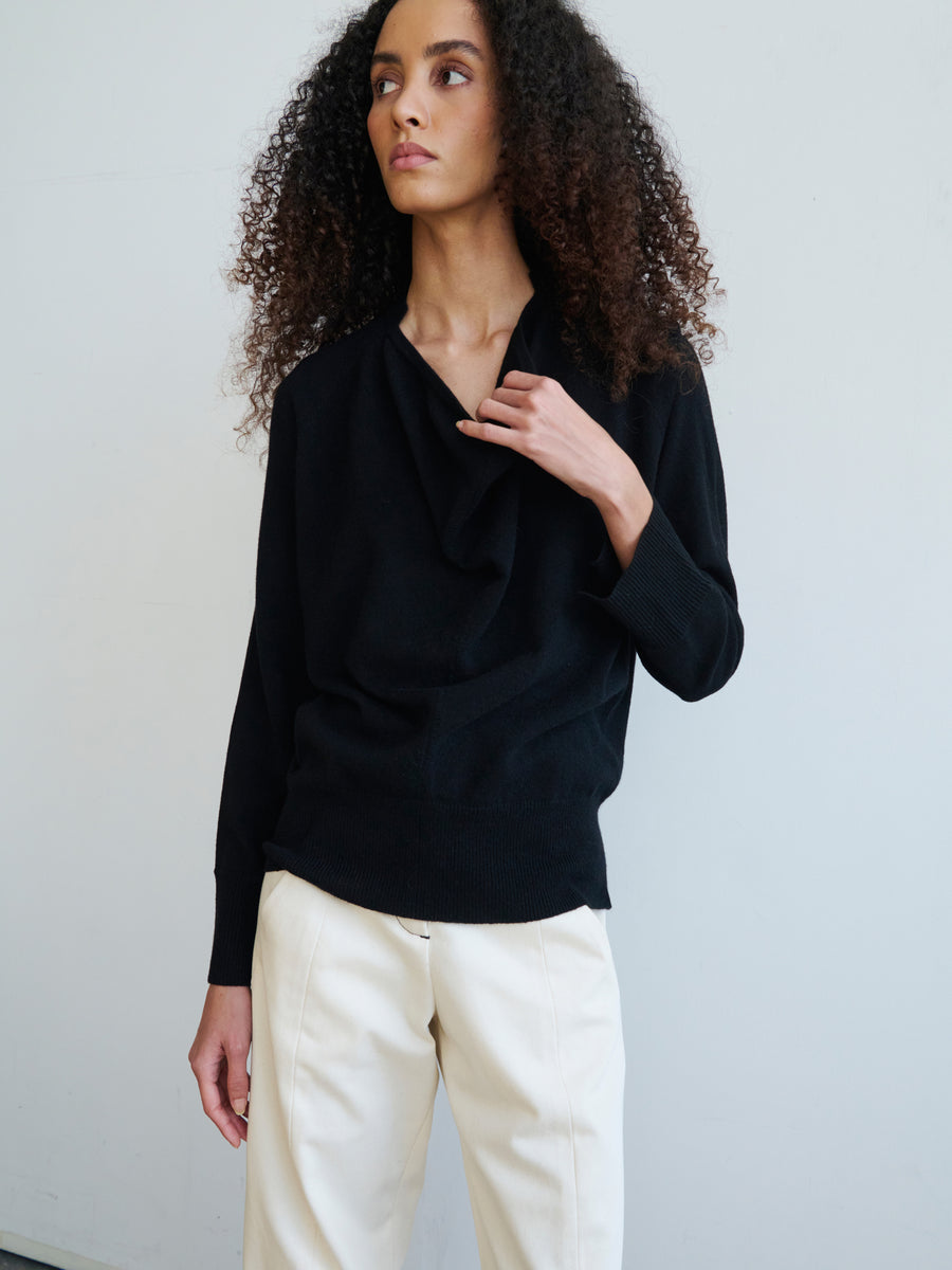 A person with curly hair is wearing the Knit Evi Top, a black, draped collar sweater by Zero + Maria Cornejo crafted from eco-friendly knitwear, paired with light-colored pants. They stand against a plain white background, gazing off to the side.