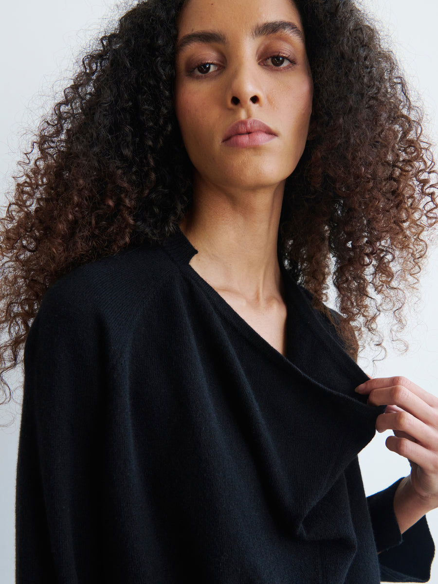 A person with curly hair is wearing the Knit Evi Top from Zero + Maria Cornejo in black, made from recycled cashmere, holding the collar with one hand. The background is a plain off-white color.