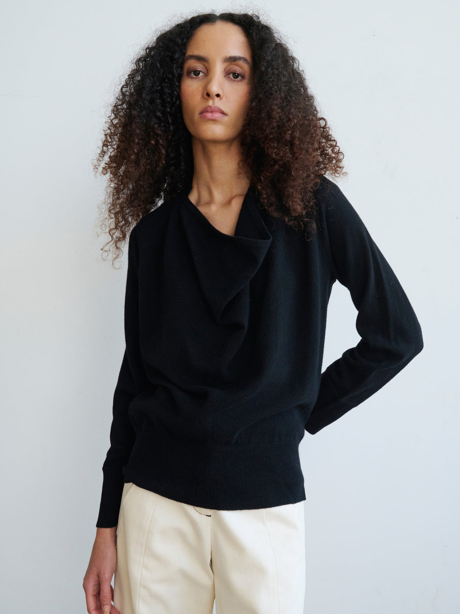 A person with curly hair is dressed in a Knit Evi Top by Zero + Maria Cornejo in black, paired with light-colored pants, standing against a plain background and gazing thoughtfully into the distance.