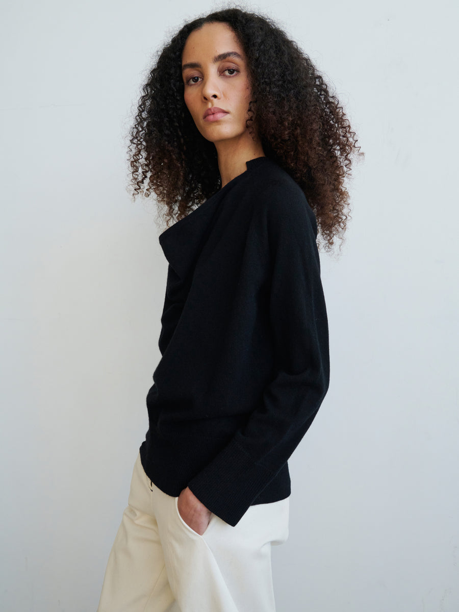 A woman with curly hair is wearing the Knit Evi Top, a black long-sleeve sweater crafted by Zero + Maria Cornejo from sustainably-sourced cashmere, paired with white pants. She stands against a plain white background, her right hand tucked into her pocket as she gazes directly at the camera.