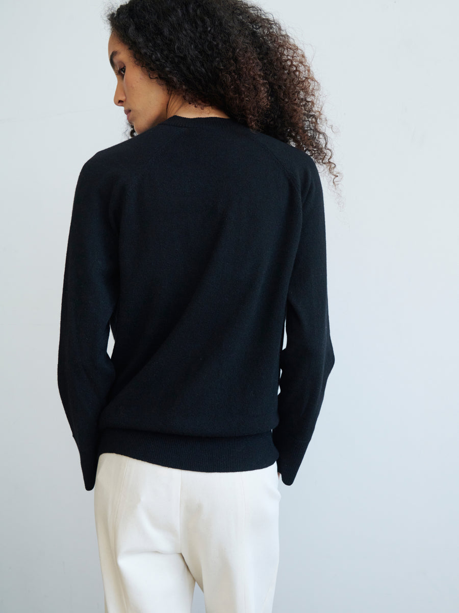 A person with curly hair is facing away from the camera, wearing the Knit Evi Top in black from Zero + Maria Cornejo and white pants. The background is plain and light-colored.