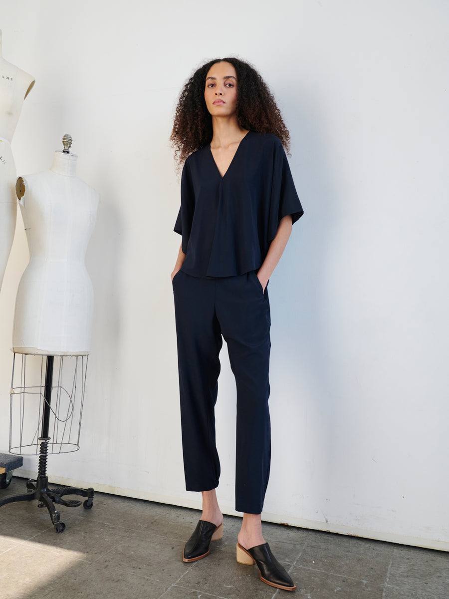 In a minimalist room, a person with curly hair exudes confidence while wearing an elegant navy blue ensemble made from stretch silk charmeuse. The V-neck top effortlessly complements the Gabi Trousers by Zero + Maria Cornejo, which feature hidden pockets. With hands tucked into the pockets, they pose confidently as a mannequin stands in the background.