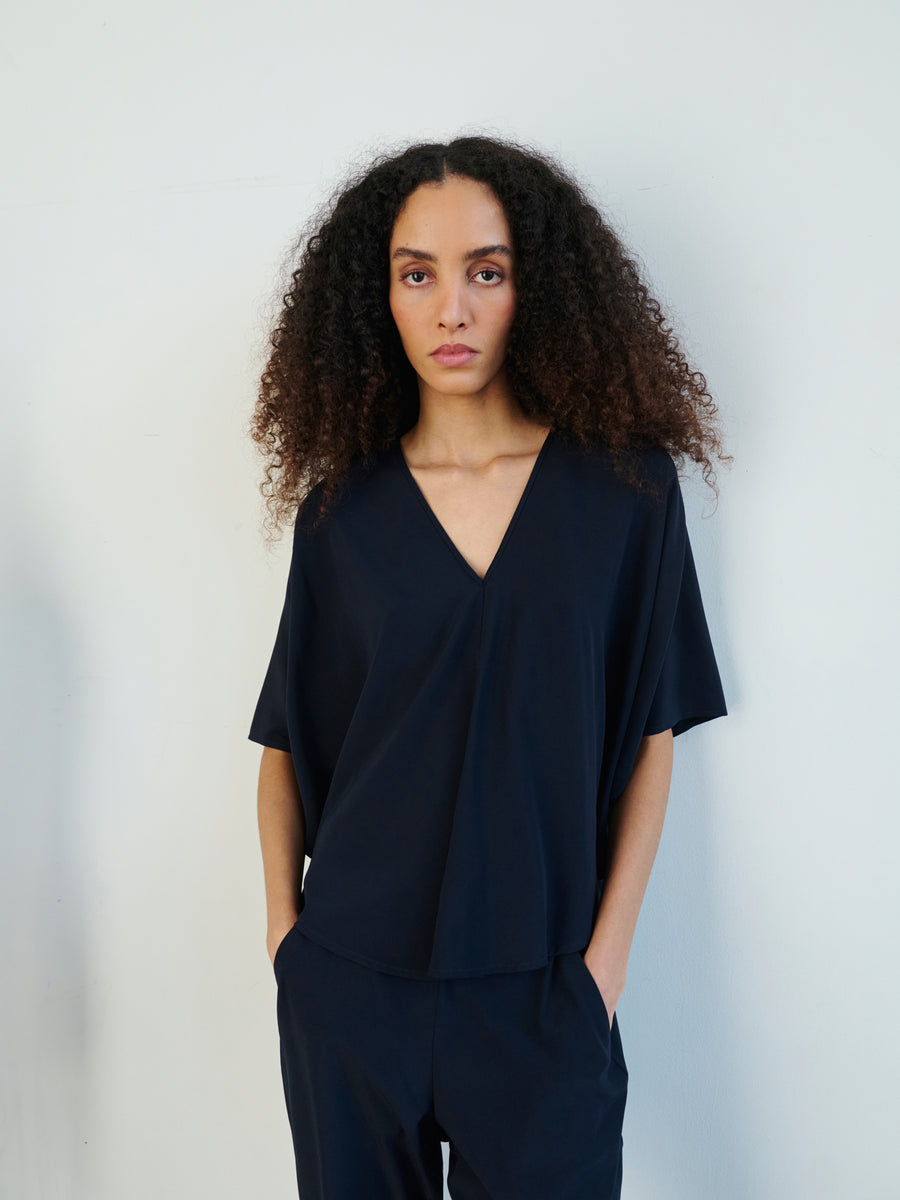 A person with curly hair stands against a plain background, wearing the Circle Top by Zero + Maria Cornejo paired with matching pants. Their hands are in their pockets, and they are looking directly at the camera.