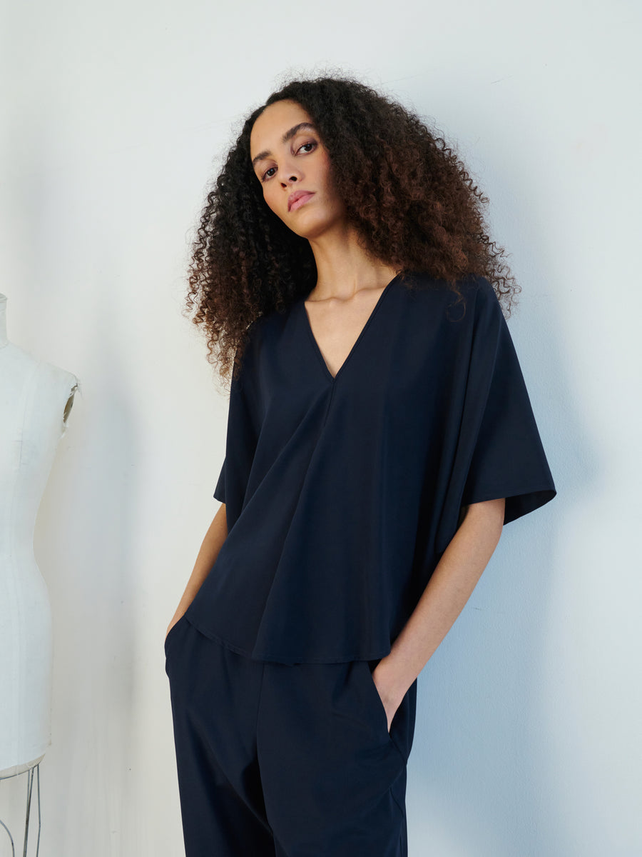 A person with curly hair is dressed in a chic, relaxed dark ensemble featuring the Circle Top by Zero + Maria Cornejo. They casually stand with their hands in their pockets against a plain background, while a mannequin displays the top's draped circle silhouette nearby.