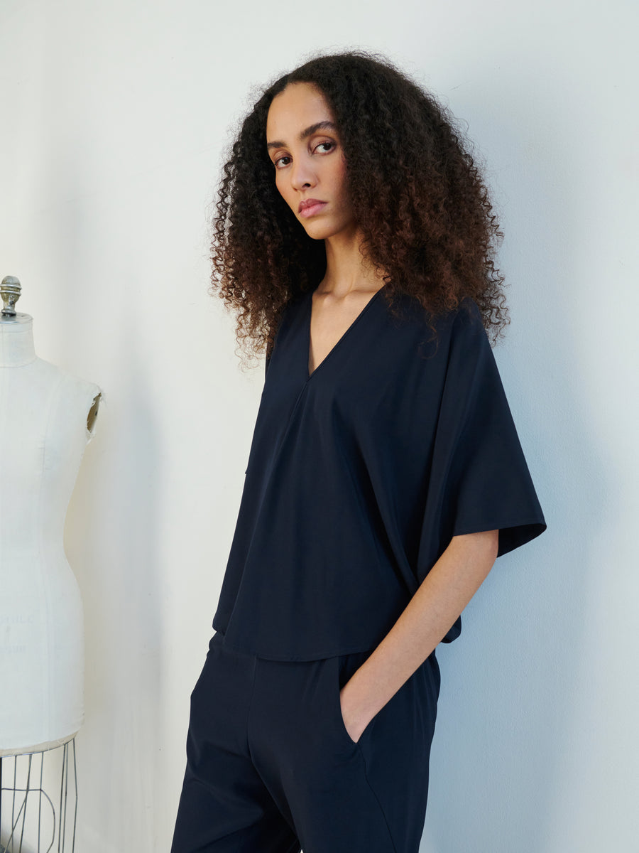 A person with curly hair stands with hands in pockets, wearing the Circle Top by Zero + Maria Cornejo, a dark blue V-neck paired with matching pants. Crafted in a draped silhouette, the ensemble exudes effortless elegance. A dress form is visible in the background.