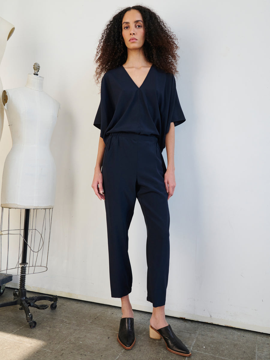 A person with long curly hair is wearing the Gabi Trouser by Zero + Maria Cornejo, paired with black mules, standing indoors next to a dress form. The outfit beautifully complements the plain white wall and concrete floor setting.