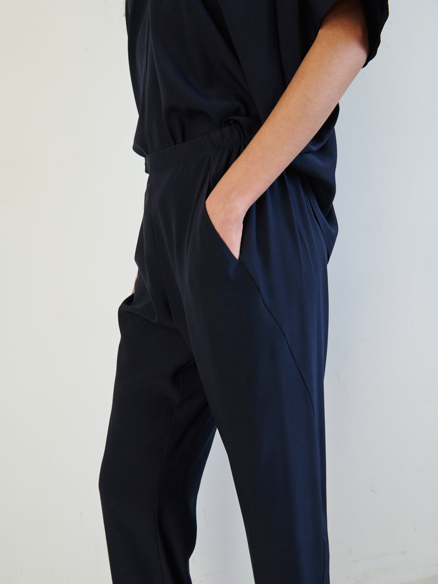 A person dressed in a relaxed, dark ensemble featuring the stretch silk charmeuse Gabi Trouser by Zero + Maria Cornejo, with their hand nonchalantly tucked into a hidden pocket. The look is completed with a long-sleeved top, set against a simple, light background.